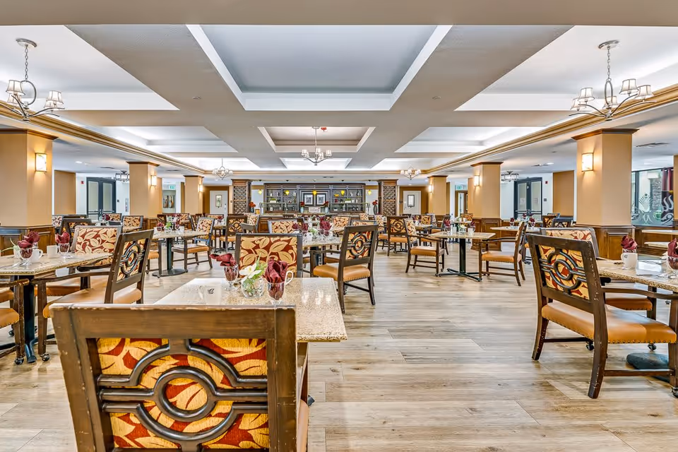 Spacious dining room with multiple tables and chairs arranged neatly. Each table has a small floral centerpiece and folded napkins. The room features wooden flooring, beige walls with wooden paneling, and a ceiling with recessed lighting and chandeliers.