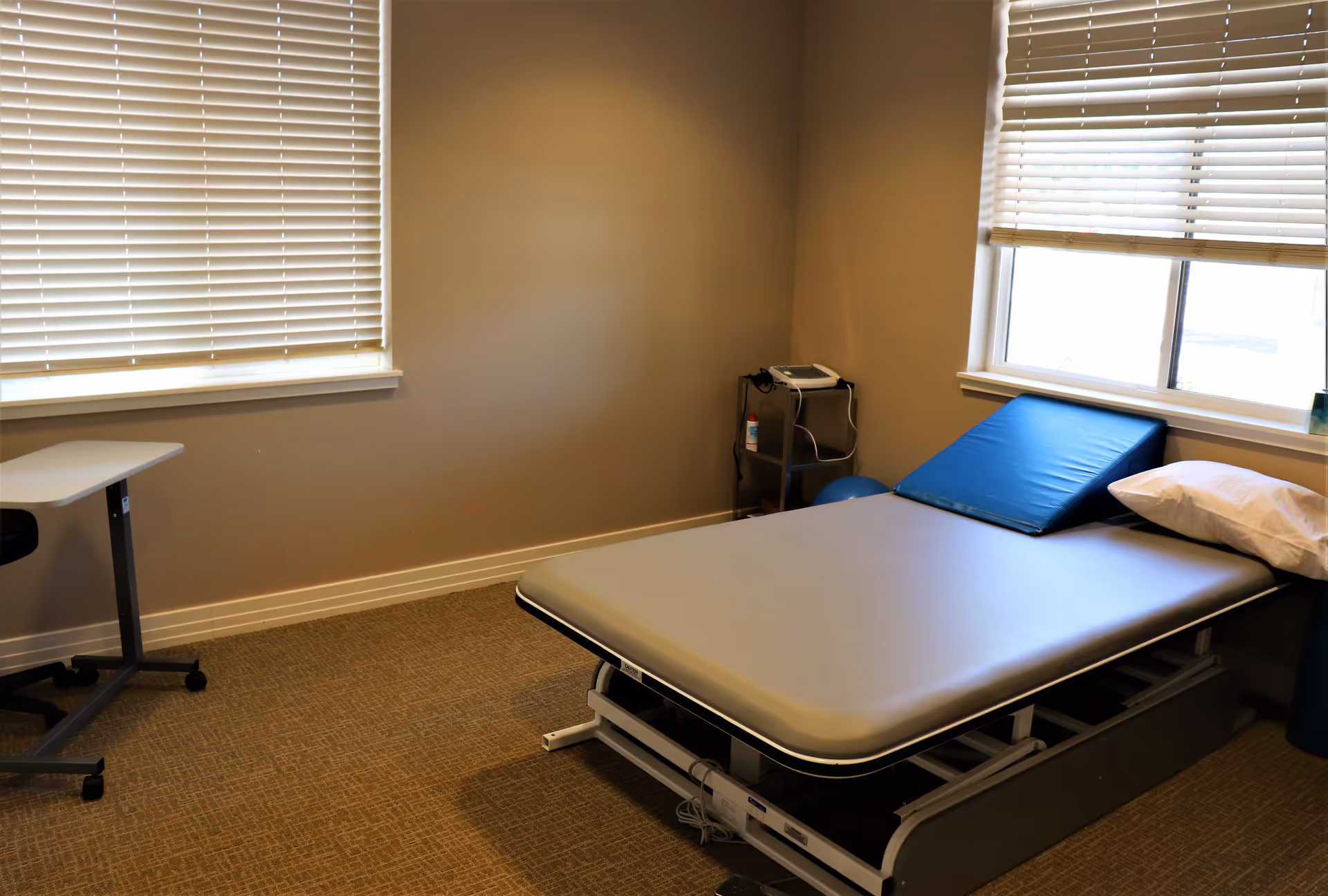 Small treatment/therapy room with an exam table, wedge cushion and pillow, windows with blinds, and a small rolling cart.
