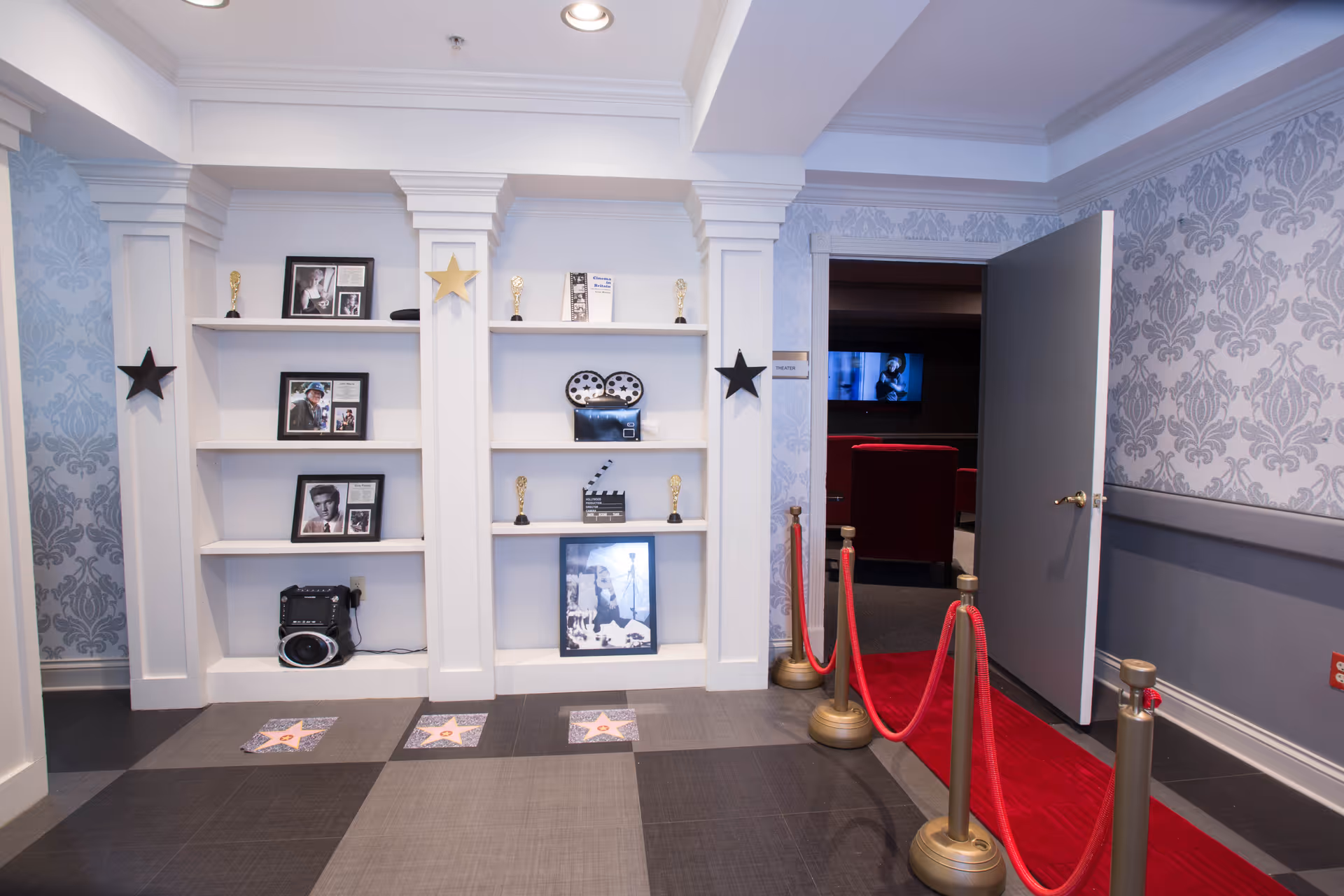 Interior view of a hallway area in an assisted living facility with white built-in shelves displaying framed photos, small trophies, and film-related decorations. The floor has star-shaped tiles resembling a walk of fame, and a red carpet with gold stanchions leads to an open door revealing a room with red theater-style chairs and a screen showing a movie scene.