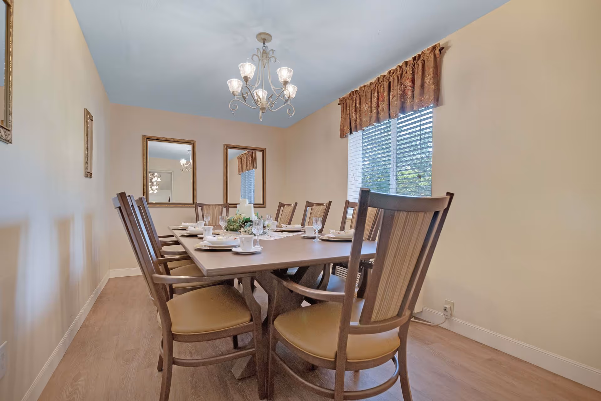 A dining room with a rectangular wooden table set for eight people with white plates, cups, and glasses. The room has beige walls, two large mirrors on one wall, a window with blinds and a brown valance, and a chandelier hanging from the ceiling.