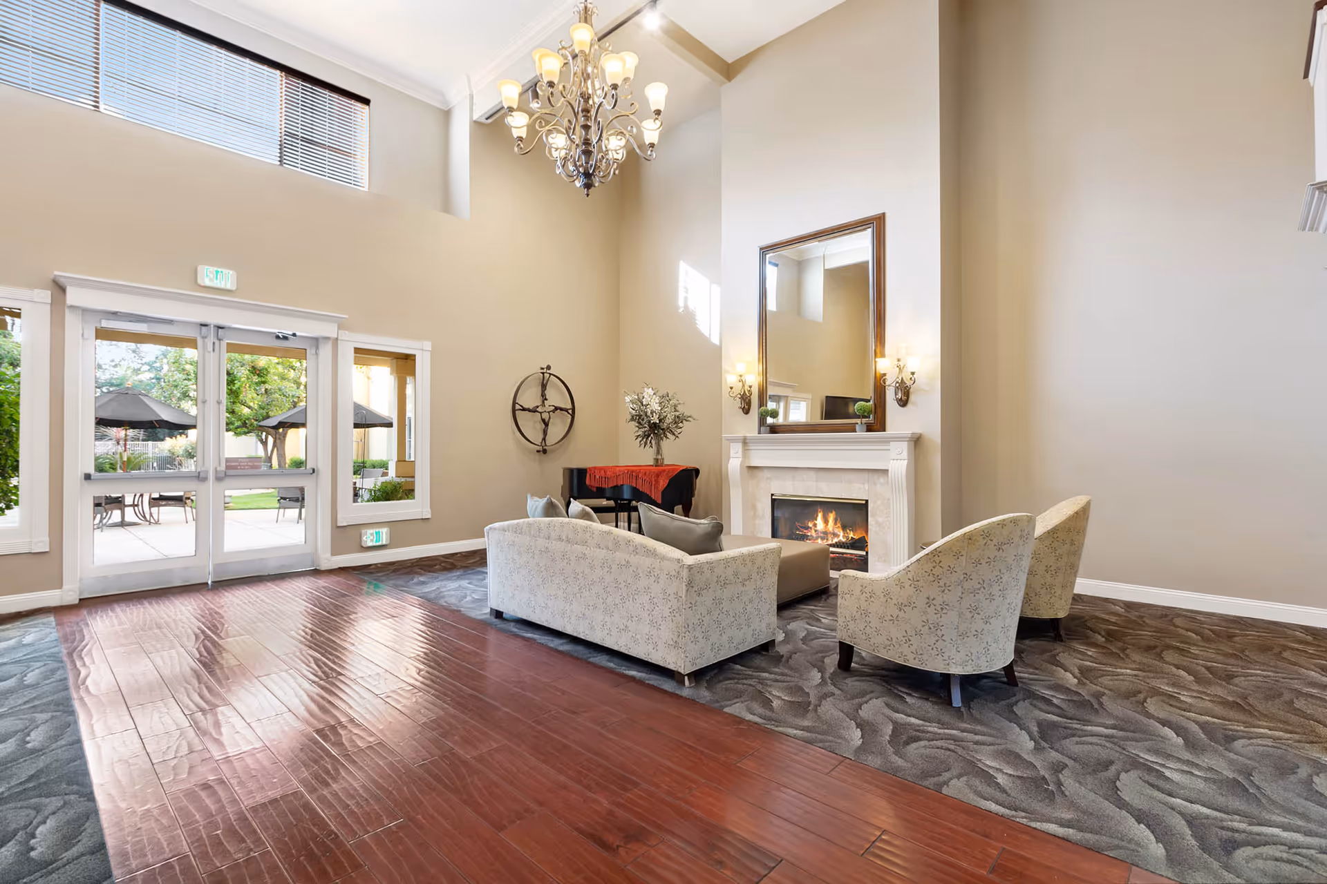 High-ceilinged lounge with seating arranged around a fireplace, a chandelier overhead, and glass doors leading to an outdoor patio.