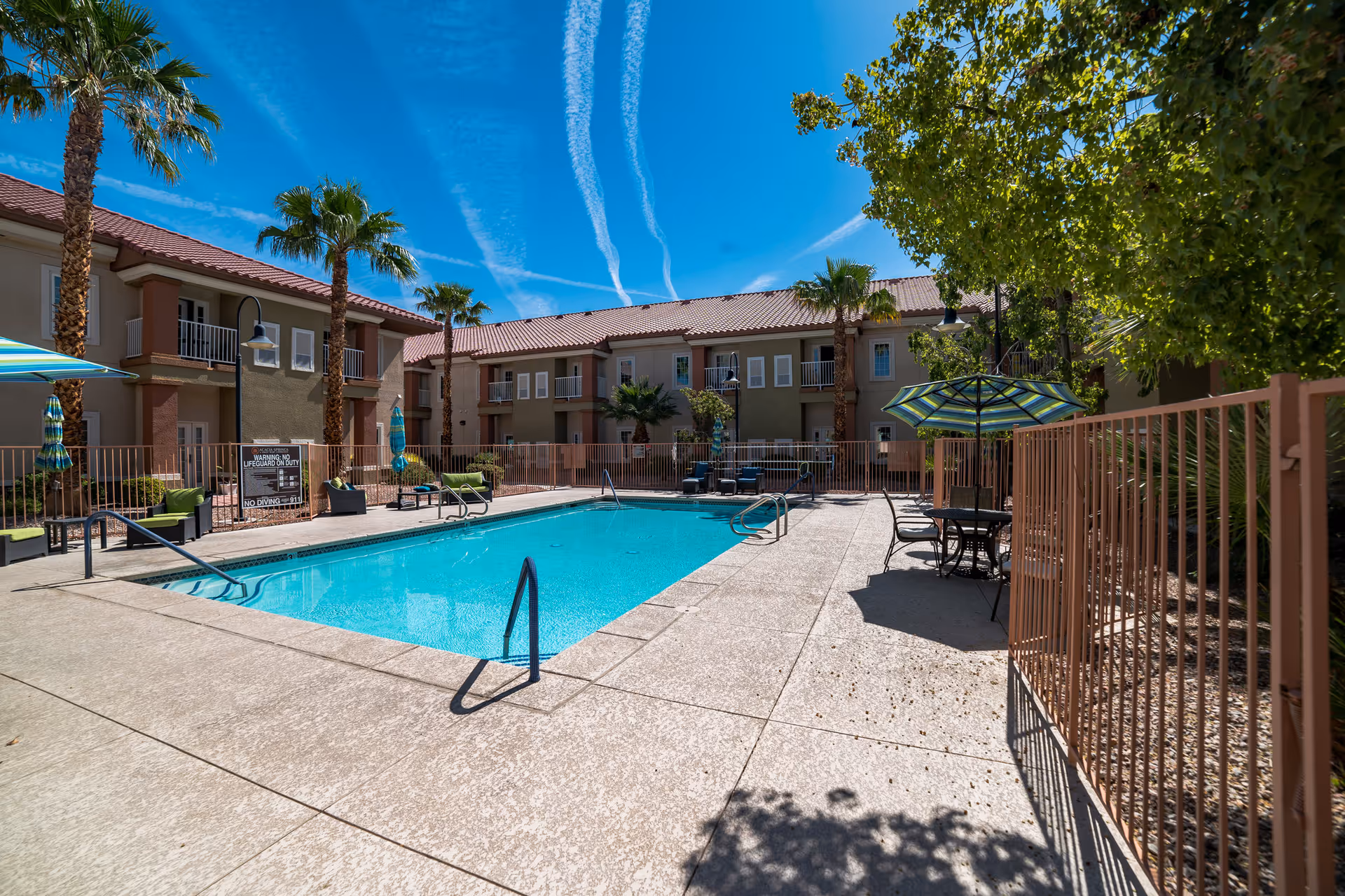 Outdoor swimming pool area at Acacia Springs facility with clear blue water, surrounded by a beige concrete deck. There are several palm trees and green plants around the pool. The pool area is enclosed by a metal fence. There are lounge chairs with green cushions and tables with striped umbrellas providing shade. The background shows a two-story building with balconies and a red-tiled roof under a bright blue sky with visible contrails.
