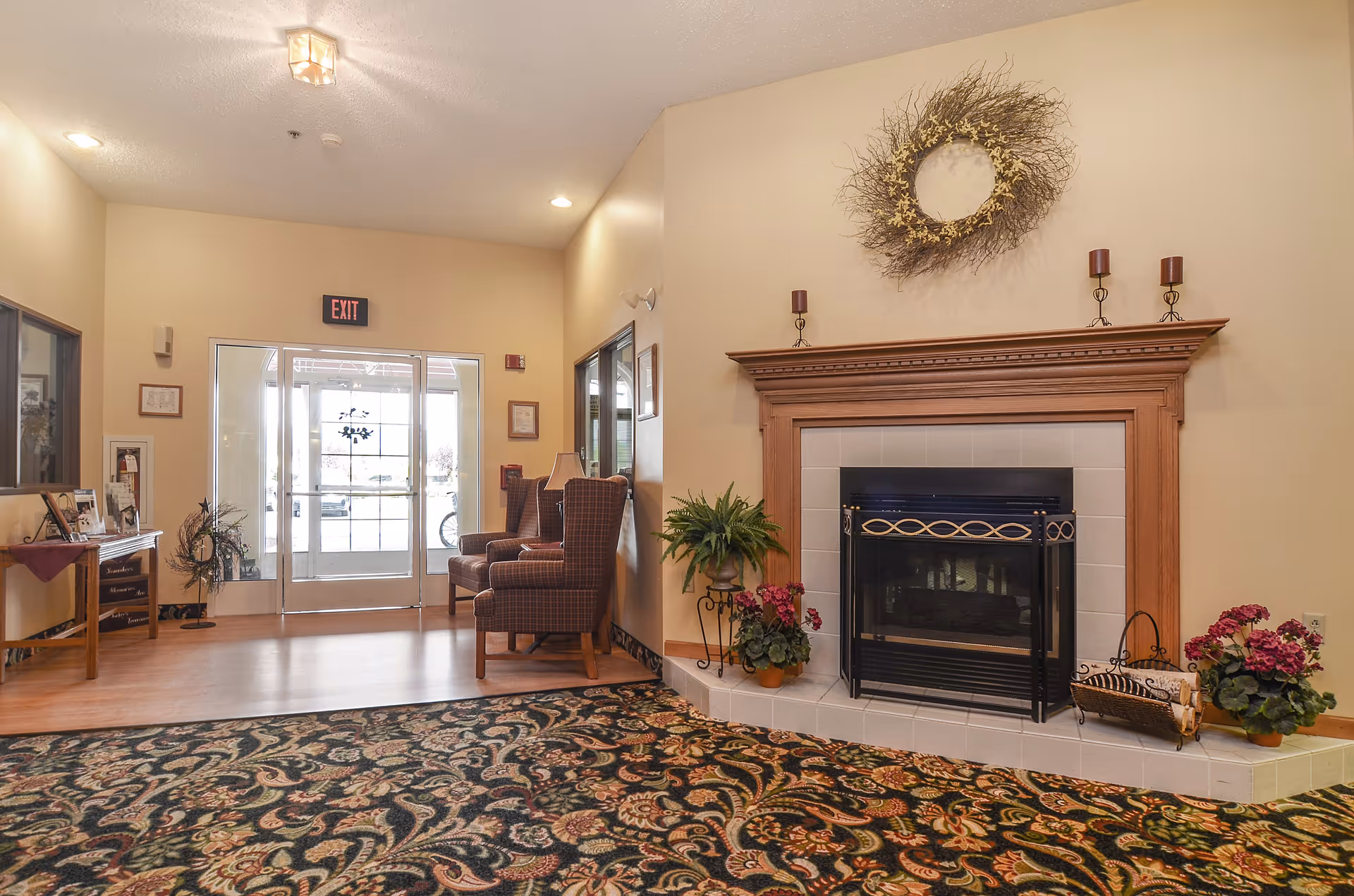Interior view of a senior living facility lobby with a fireplace on the right decorated with plants, candles, and a wreath above it. Two upholstered armchairs are placed near the glass entrance doors with an exit sign above. A patterned carpet covers the floor in the foreground, and a small table with framed photos and decorations is on the left side.