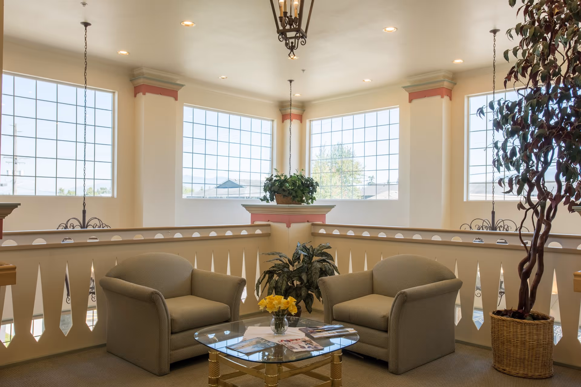 Two armchairs and a glass coffee table in a bright upstairs lounge with large grid windows and potted plants.
