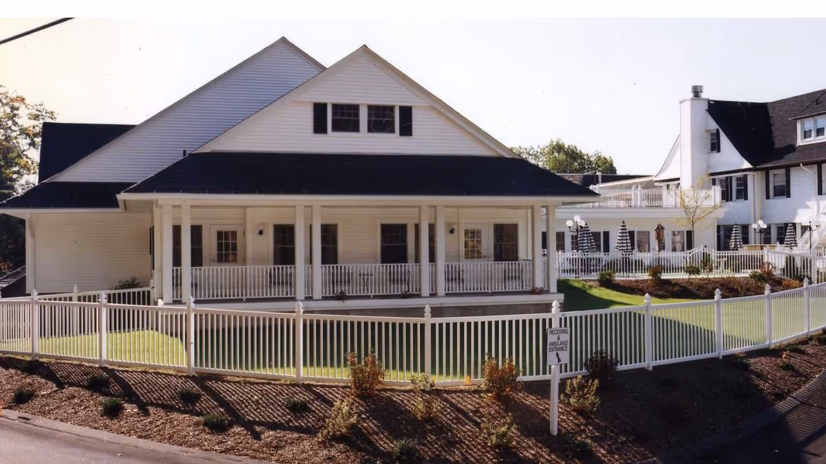 White multi-story building with a covered front porch, white picket fence, and manicured lawn.