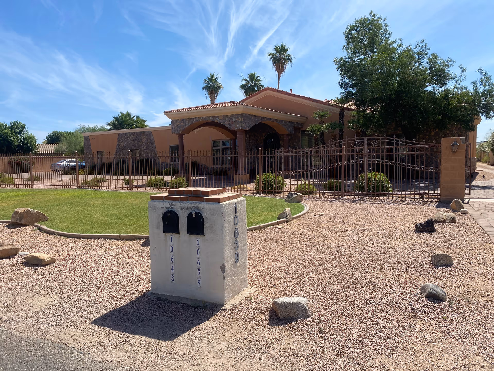 Front exterior of a single-story assisted living building with a gated entrance, mailbox pillar, and desert landscaping under a blue sky.