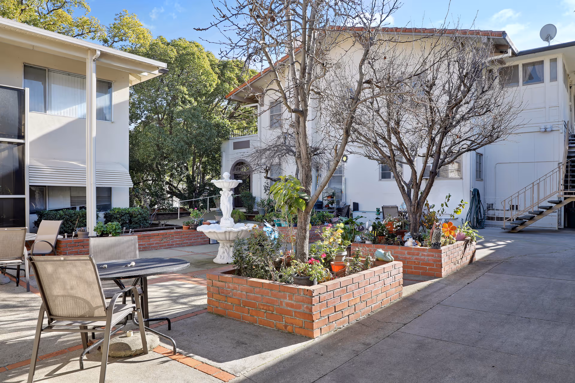 Outdoor courtyard area of Ivy Ridge Assisted Living featuring brick planter boxes with trees and plants, a white decorative fountain, patio tables and chairs, and surrounding buildings under a clear blue sky.