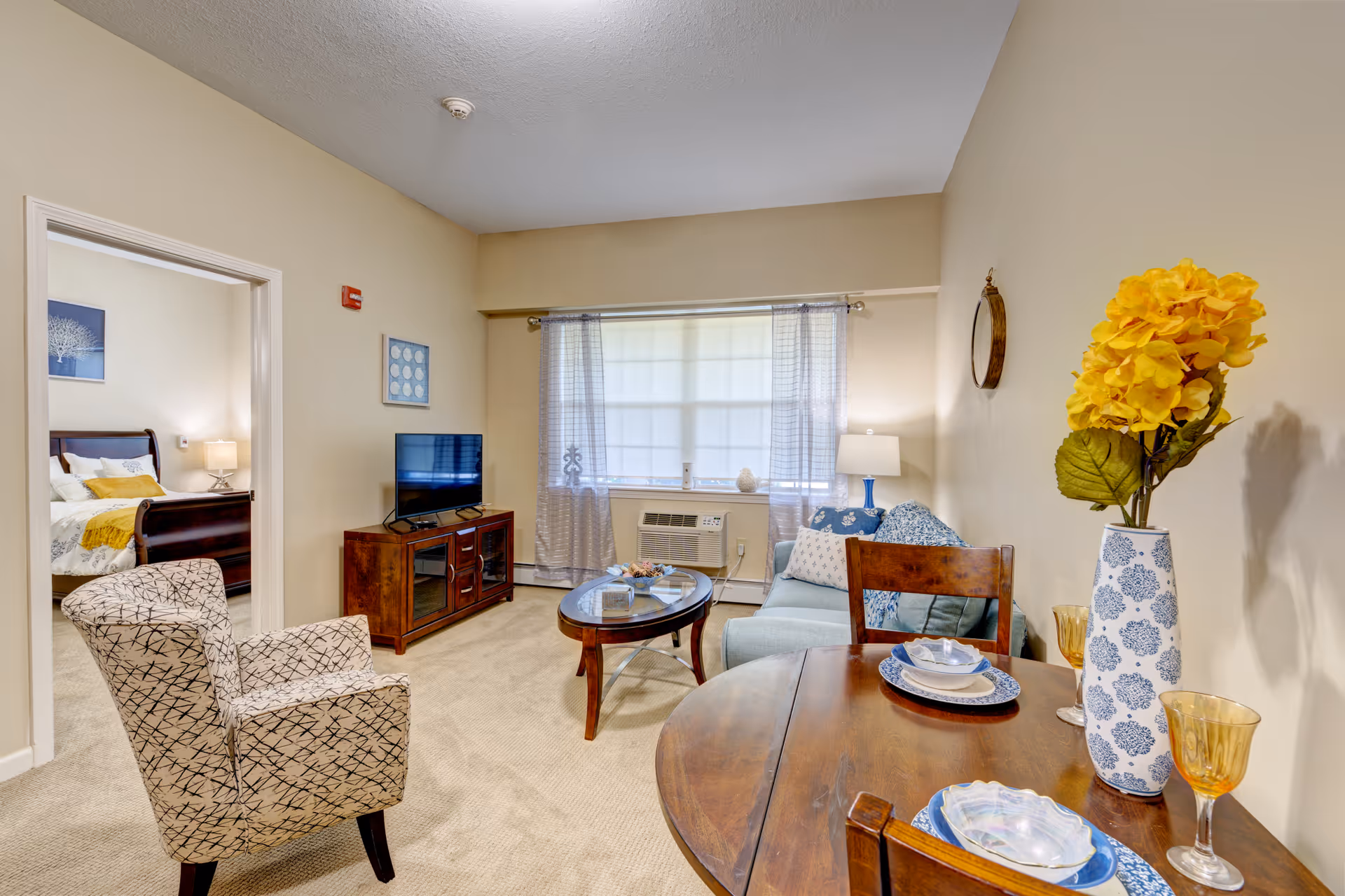 A cozy living room area with a patterned armchair, a wooden coffee table, a small blue sofa with cushions, and a TV on a wooden stand. A window with sheer curtains and an air conditioning unit is visible. To the right, a round wooden dining table is set with plates, bowls, and amber-colored glasses, and a vase with yellow flowers. A bedroom with a bed and nightstand is visible through an open doorway.