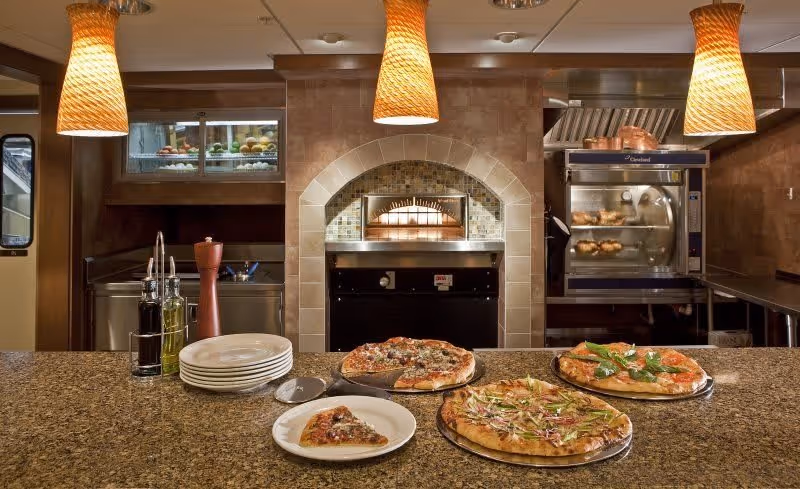A kitchen counter with three pizzas and a slice on a white plate in the foreground. Behind the counter are kitchen appliances including a pizza oven with a fire inside, a rotisserie oven with chickens, and hanging pendant lights above the counter.