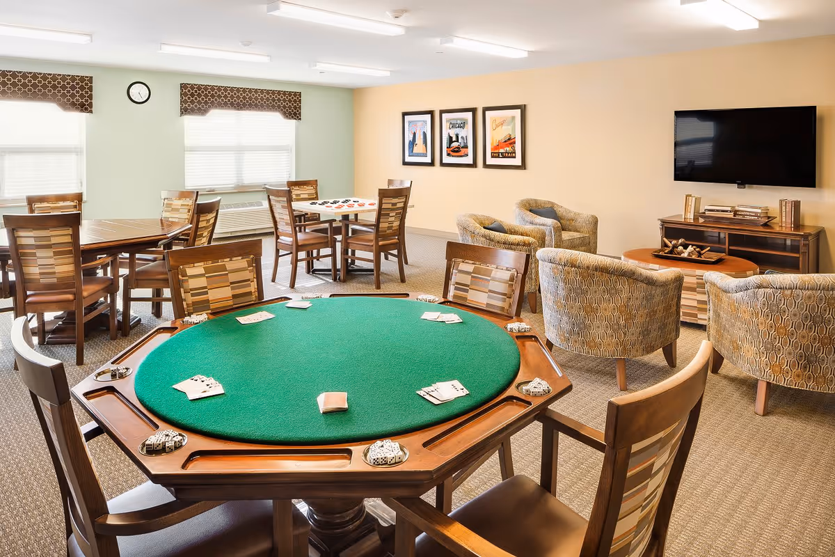 A well-lit common room with a green felt poker table in the foreground, surrounded by wooden chairs. In the background, there are additional tables and chairs, a wall-mounted TV, three patterned armchairs, and framed artwork on the wall. The room has windows with patterned valances and a clock above one window.