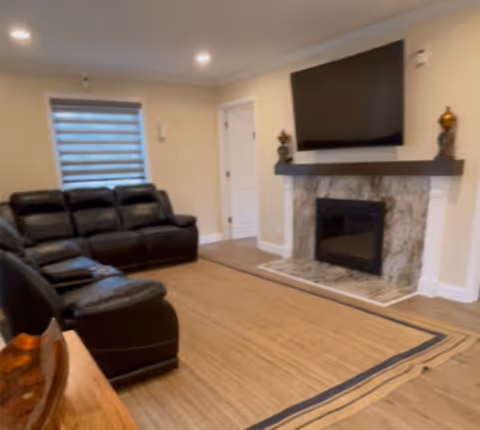 A living room with a black leather sectional sofa, a large flat-screen TV mounted above a marble fireplace, beige walls, wooden flooring, and a window with a modern horizontal blind.