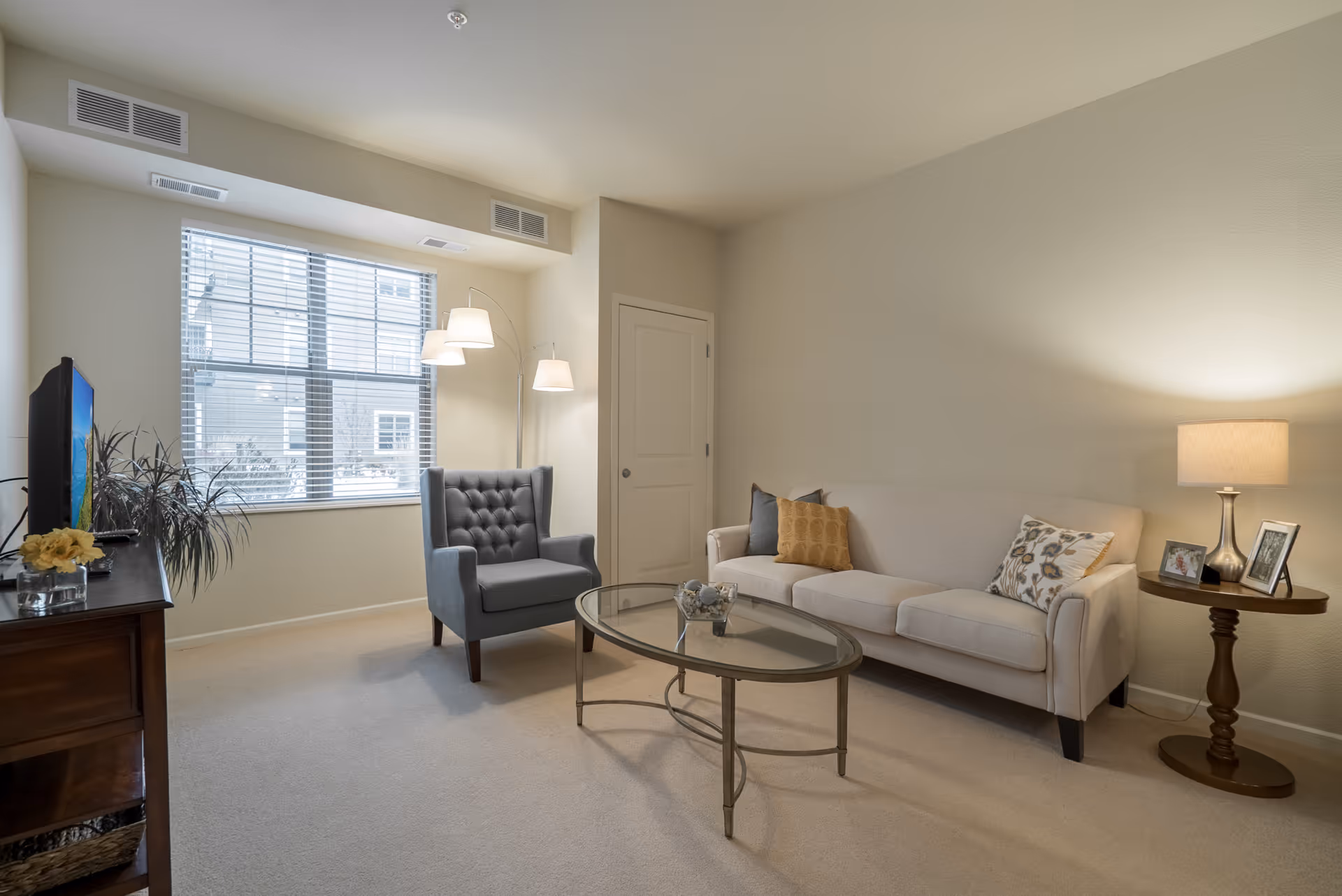 A cozy living room with a beige sofa adorned with three decorative pillows, a gray armchair, and a glass coffee table. A wooden side table with a lamp and framed photos is next to the sofa. A window with blinds allows natural light to brighten the room. A TV sits on a wooden cabinet with a plant and a small flower arrangement.
