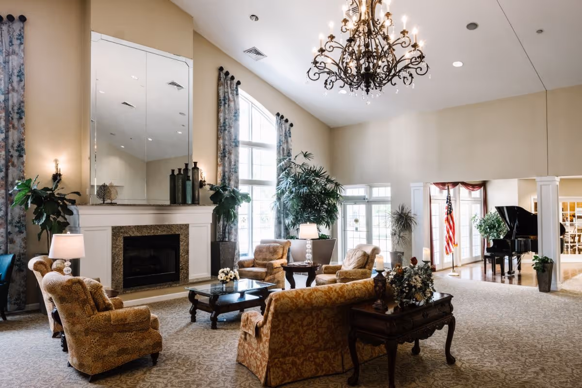 A spacious and elegant living room area with high ceilings, a large chandelier, and tall windows with floral curtains. The room features a fireplace with a large mirror above it, several upholstered armchairs and sofas arranged around a glass coffee table, potted plants, and a grand piano near the entrance with an American flag beside it.