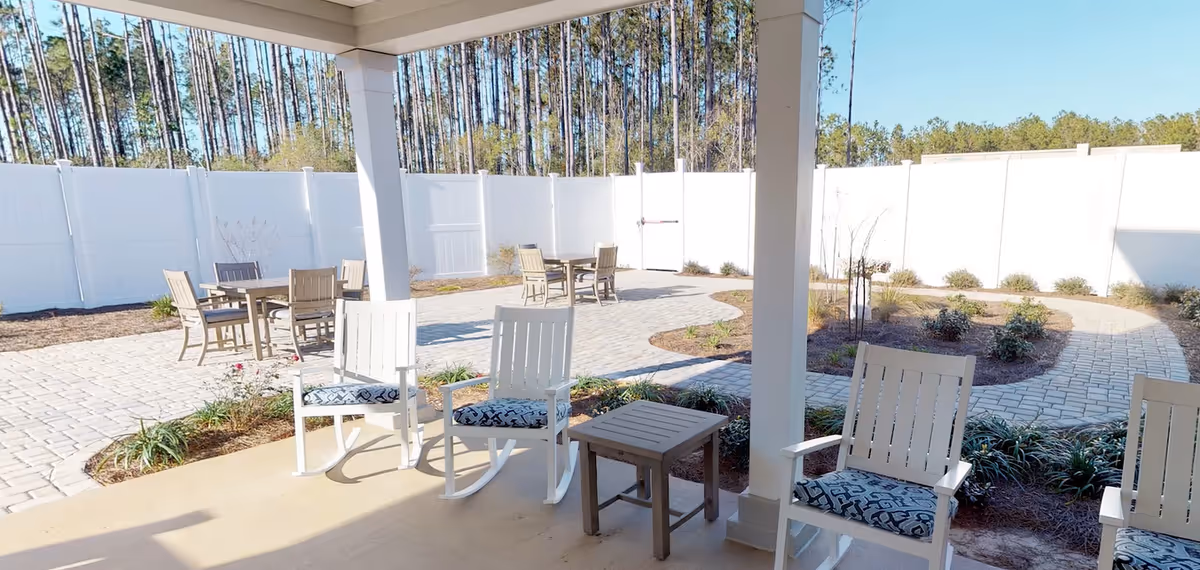 Outdoor patio area with white rocking chairs and a small table under a covered porch, overlooking a paved walkway and garden beds surrounded by a white privacy fence and tall trees in the background.