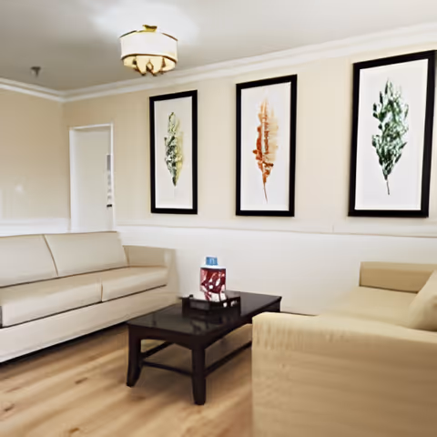 A living room with two beige sofas facing each other, a dark wooden coffee table in the center with decorative items on it, and three framed leaf artworks hanging on the wall behind one sofa. The room has light-colored walls, wooden flooring, and a ceiling light fixture.