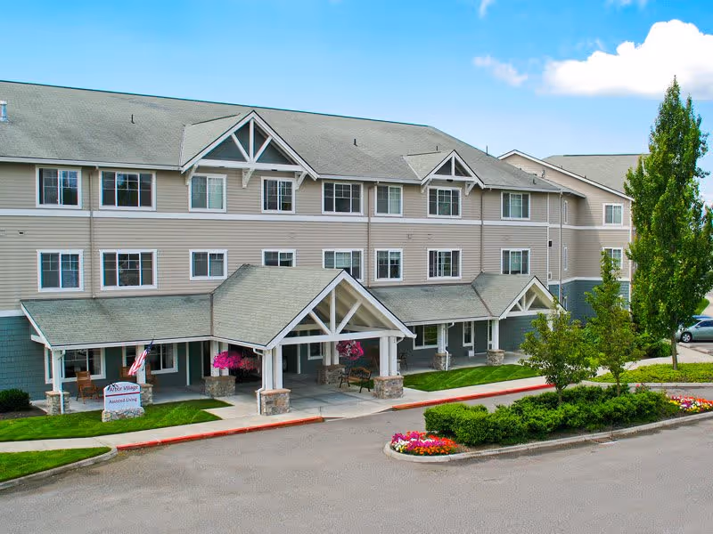 Exterior view of a three-story senior living facility building with beige siding and a gray roof. The entrance features a covered porch with white pillars and hanging flower baskets. There is a small landscaped area with green bushes and colorful flowers near the driveway. A sign near the entrance reads 'Arbor Village Assisted Living.'