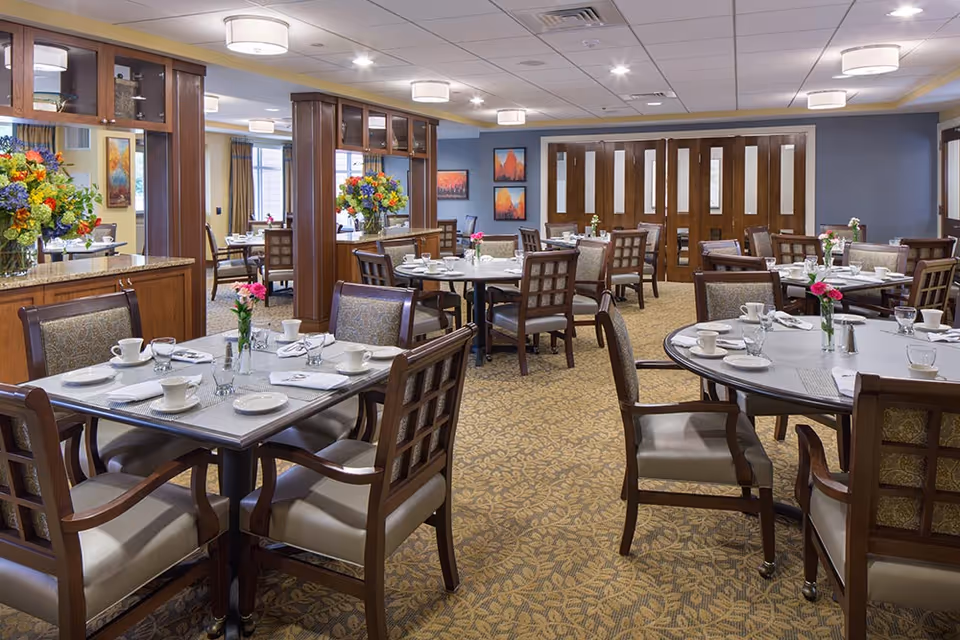 A spacious dining room in a senior living facility with multiple round and square tables set with white plates, cups, glasses, and napkins. Each table has a small vase with pink flowers. The room features wooden chairs with cushioned seats, patterned carpet, and decorative wooden columns with glass shelving. There are colorful flower arrangements on the counters and framed artwork on the walls.