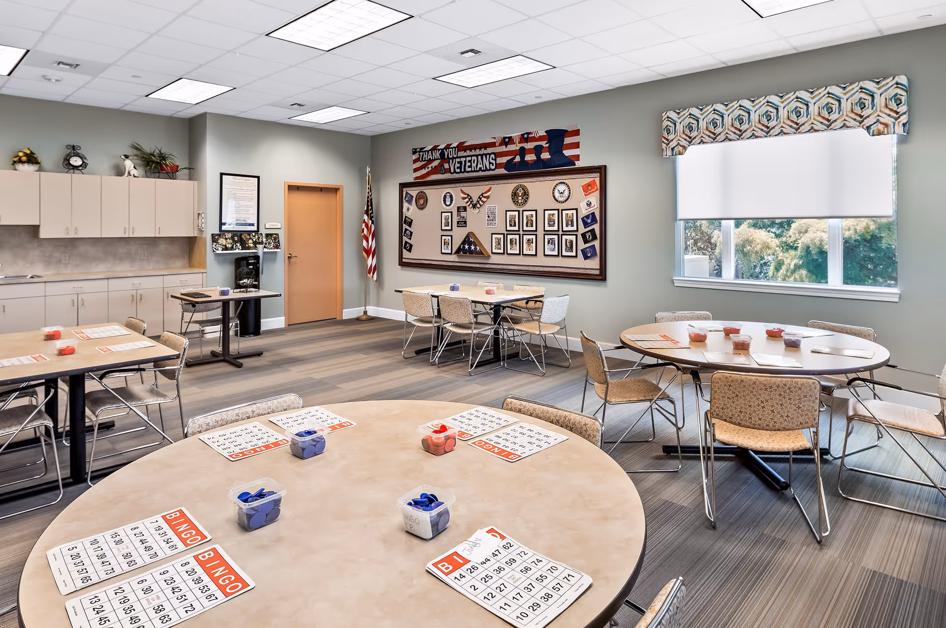 A bright activity room with round tables set up for bingo, each table having bingo cards and containers with red and blue bingo chips. The room has beige walls, a large window with a patterned valance, and a bulletin board displaying a 'Thank You Veterans' banner along with various photos and memorabilia. There are chairs around the tables and cabinets along one wall.