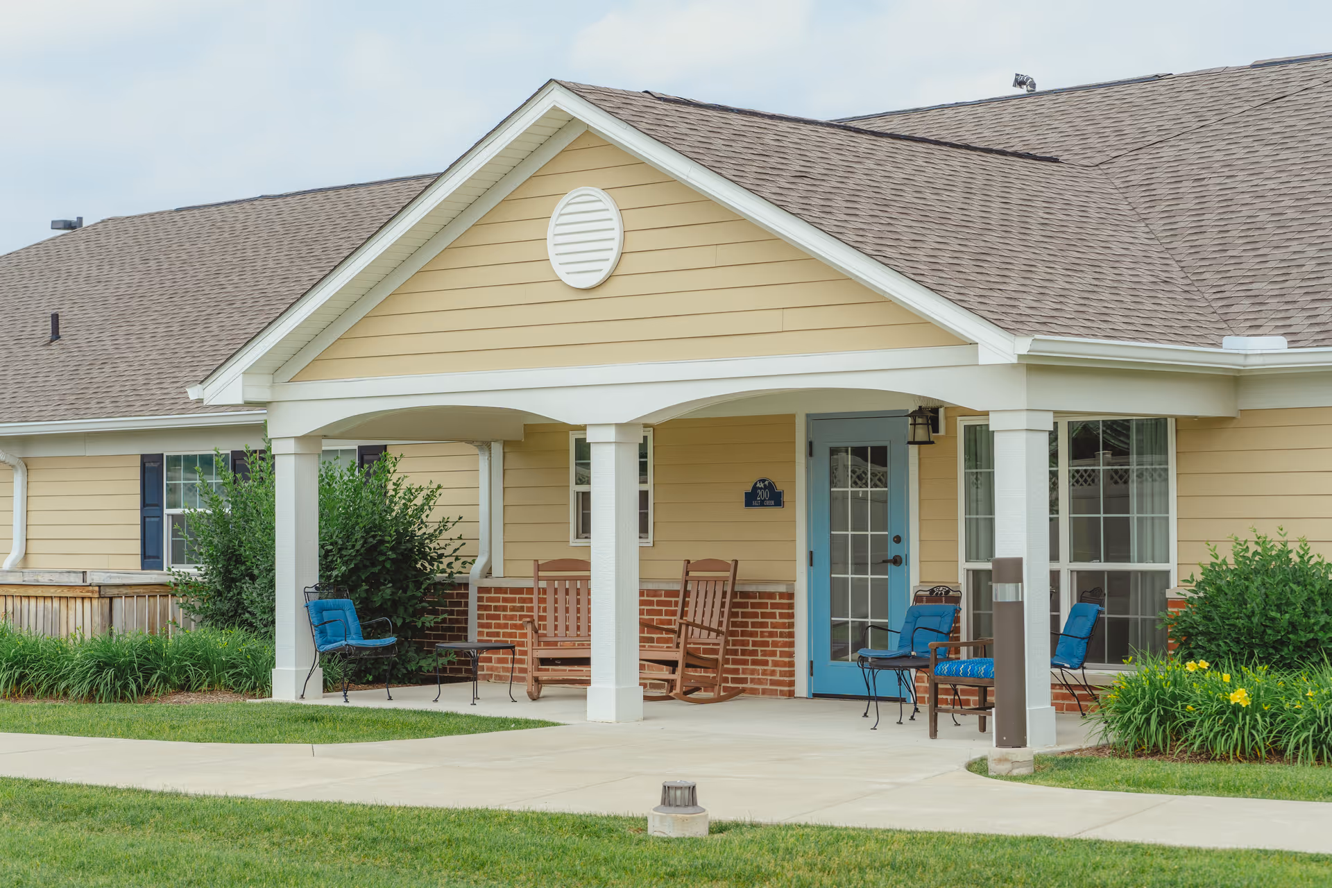 Covered porch area of a residential building with beige siding and a blue door. The porch has white columns and is furnished with wooden rocking chairs and metal chairs with blue cushions. There are green bushes and yellow flowers near the porch, and a concrete walkway in front.
