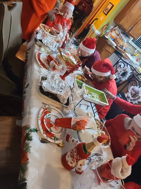 A festive holiday meal scene with several people wearing Santa hats seated around a dining table covered with a white tablecloth decorated with holly. The table is set with holiday-themed plates, red cups, and various dishes including guacamole and other foods. The background shows a kitchen area with wooden cabinets and holiday decorations.