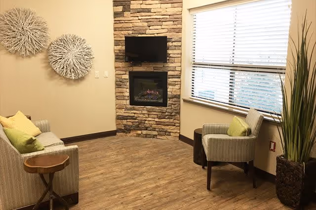 A cozy living room area with a stone fireplace and a mounted flat-screen TV above it. The room has wooden flooring, a large window with blinds, two armchairs with green cushions, a small wooden side table, and decorative wall art made of circular woven materials. There is also a tall plant in a pot near the window.
