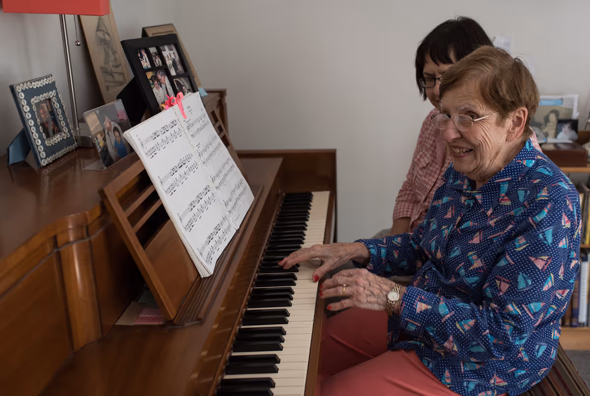 An elderly woman smiling and playing the piano with the assistance of another woman sitting beside her in a cozy room with framed photos on the piano and shelves in the background.