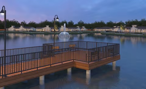Wooden pier with benches and lampposts extending over a pond with a fountain and Wilshire Senior Living buildings along the shore at dusk.