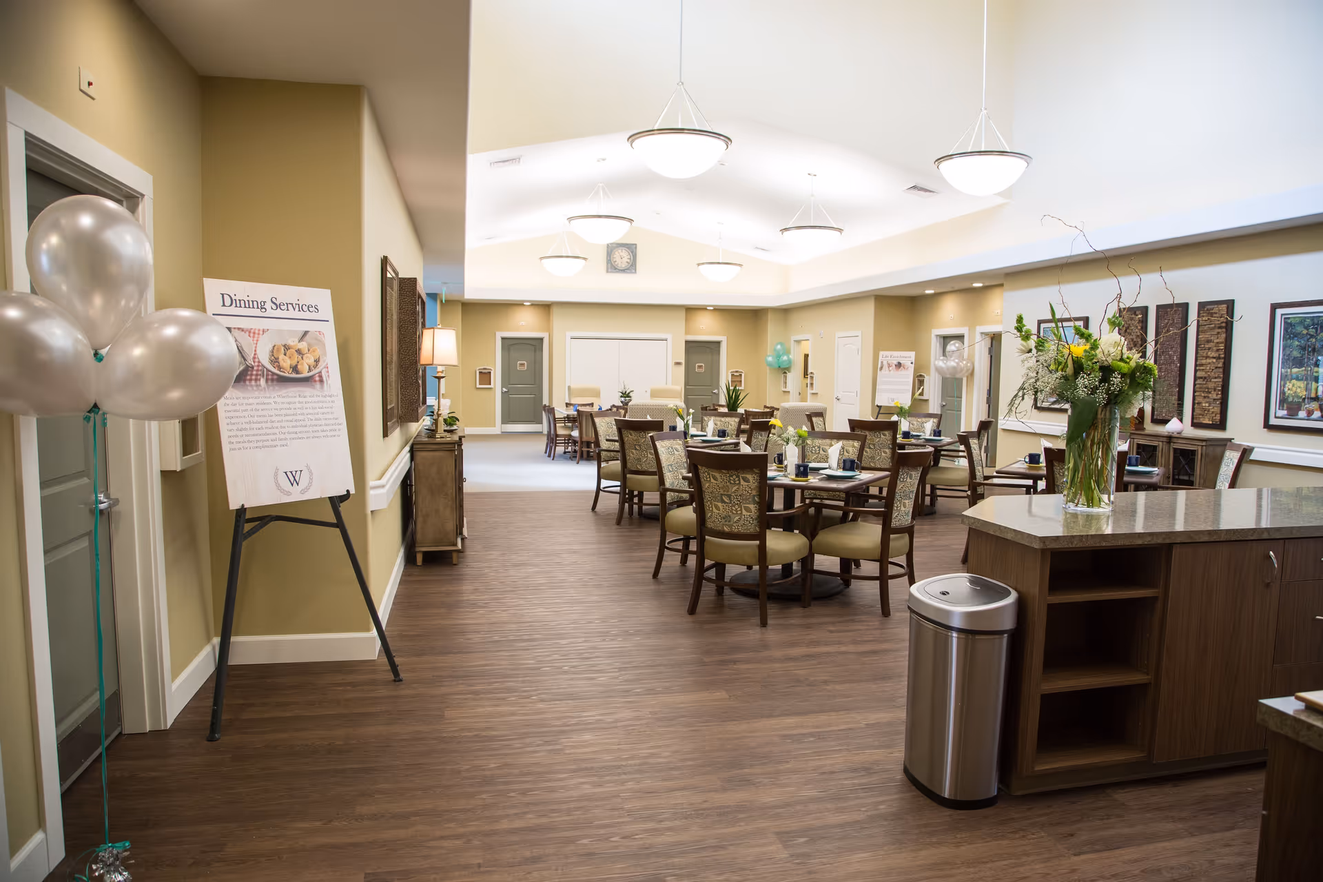 A spacious dining room in a senior living facility with several round tables and chairs arranged neatly. The room has warm beige walls, wooden flooring, and modern hanging light fixtures. There is a sign on an easel labeled 'Dining Services' near the entrance, along with silver balloons. A countertop with a vase of flowers and a trash can is visible on the right side. The room is well-lit with natural and artificial light.