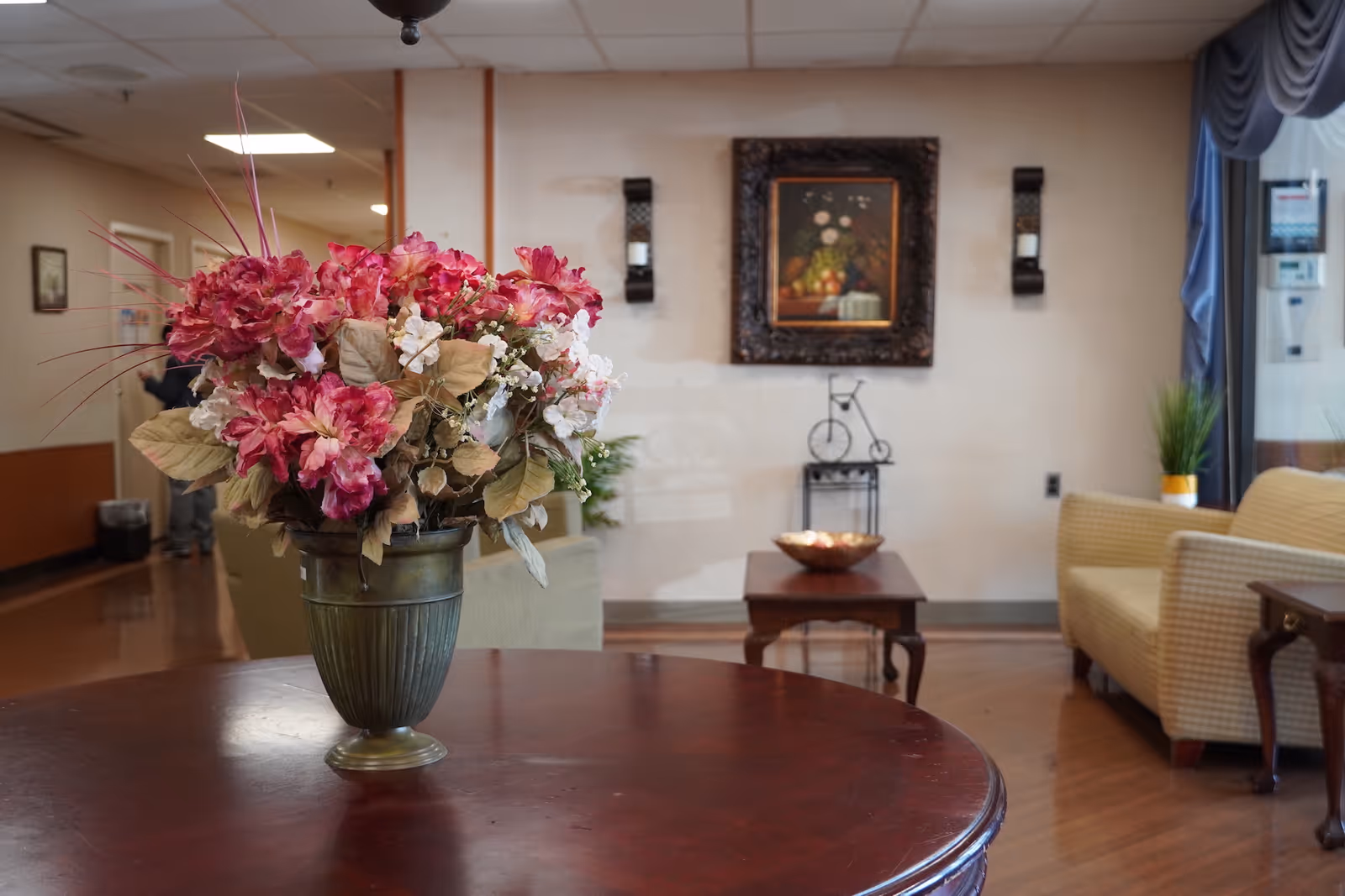 A vase of pink and white flowers sits on a round wooden table in a cozy lounge area with armchairs, a painting, and side tables.