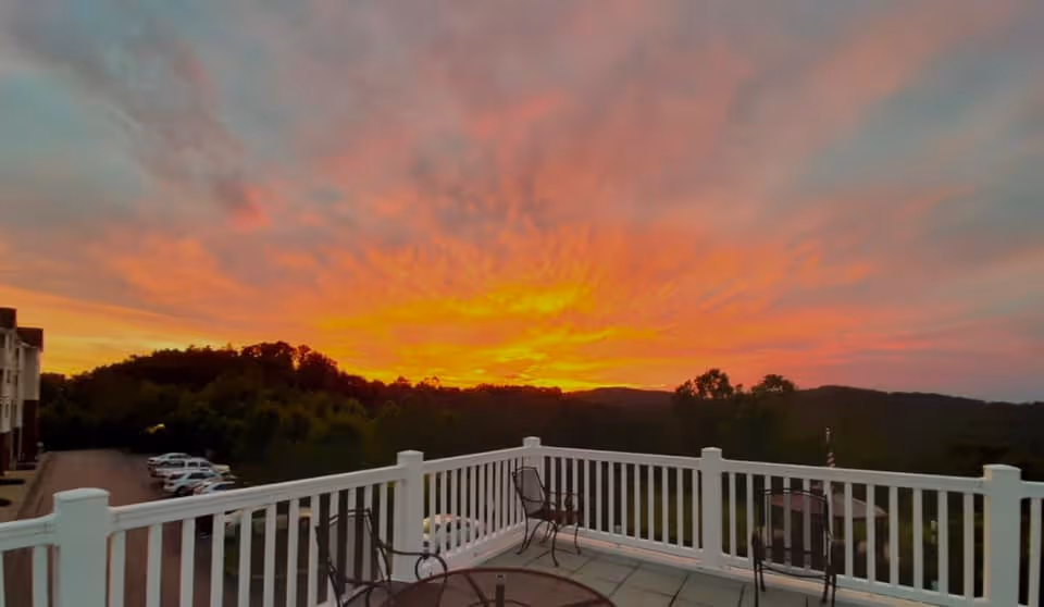 View of a vibrant sunset with orange and pink hues over distant hills, seen from a balcony with white railings and outdoor chairs. A parking lot with cars and part of a building are visible to the left.