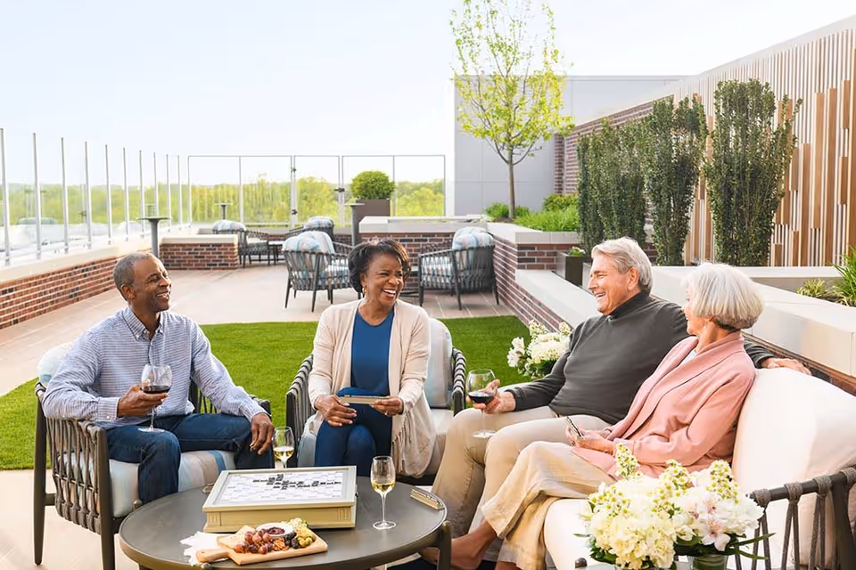 Four older adults sitting and enjoying drinks while playing a board game on a rooftop patio with greenery and comfortable seating.