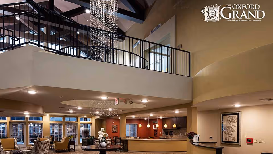 Spacious senior living facility lobby with seating, a chandelier, upper balcony, and reception desk.