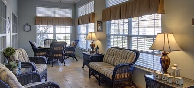 A sunlit sitting area with wicker furniture including two armchairs and a loveseat with striped cushions, a small round table with four wicker chairs near large windows with blinds and valances, two table lamps on side tables, and decorative items including a clock and a block sign spelling 'laugh'.