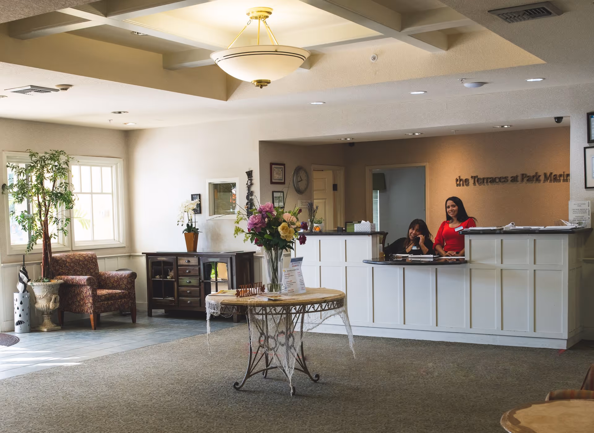 Reception area of The Terraces at Park Marino with two staff members behind a white front desk. The room has a round table with a flower arrangement in the center, a patterned armchair near a window, a potted plant, and a side cabinet. The lighting is warm and the space appears welcoming.