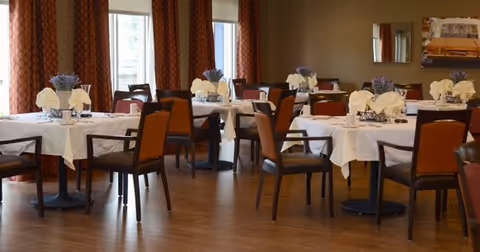 Dining room with multiple round tables set with white tablecloths, napkins and floral centerpieces and brown chairs.