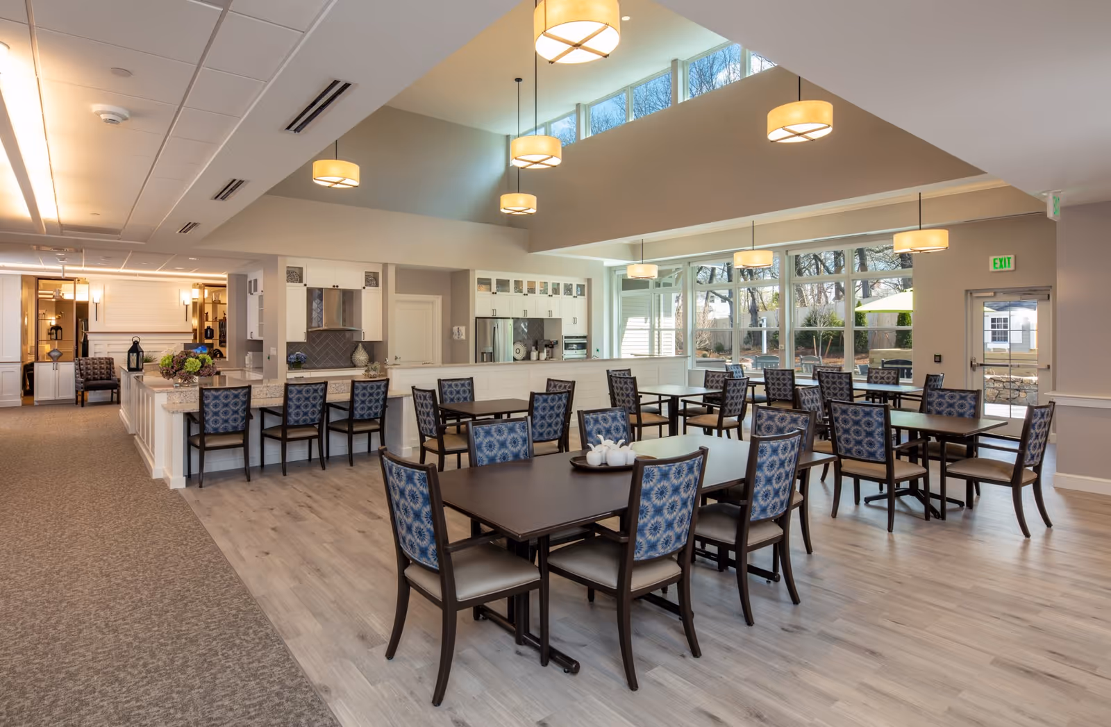 A spacious and well-lit dining area in a senior living facility featuring multiple dark wood tables with blue patterned chairs. The room has large windows allowing natural light to fill the space, modern pendant lights hanging from the ceiling, and a kitchen area with a countertop and bar stools in the background. The flooring is a combination of light wood and carpet, and there is an exit door leading outside.