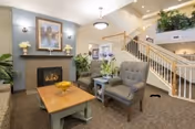 A cozy senior living facility common area featuring a fireplace with a framed painting above it, two upholstered armchairs, a wooden coffee table with a decorative bowl, and a side table with a flower arrangement. A staircase with wooden railings leads to an upper floor, and there are plants and soft lighting throughout the space.