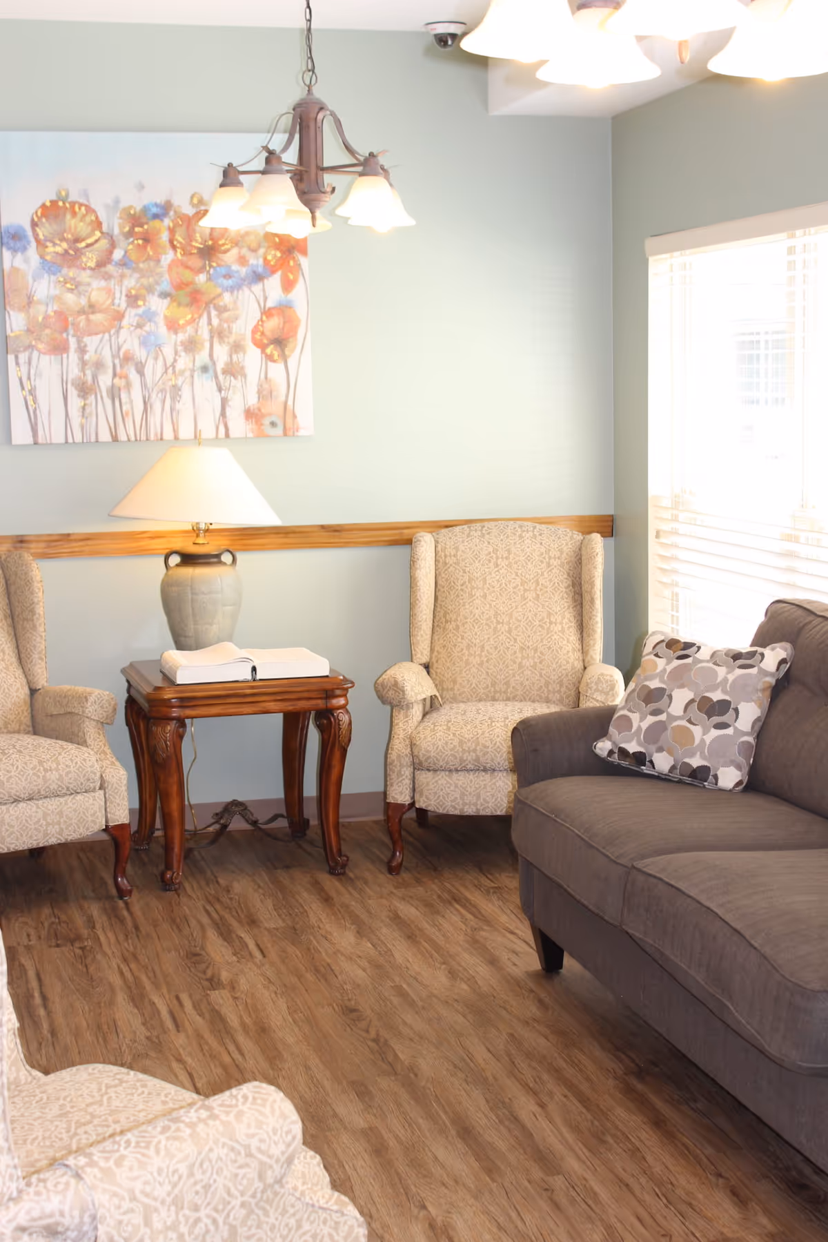 A bright sitting room with two upholstered wingback chairs, a gray sofa, a wooden side table with a lamp, and floral wall art.