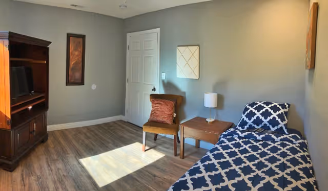 A small bedroom with a single bed covered in a blue and white patterned blanket, a wooden nightstand with a small lamp, a brown chair with a red cushion, a wooden cabinet with a TV, and two framed wall decorations. The room has wooden flooring and light gray walls with a closed white door.
