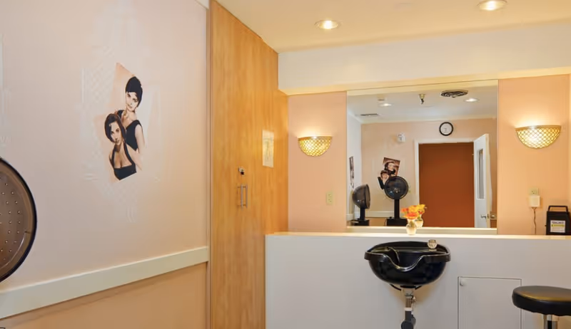 Interior view of a hair salon area in a skilled nursing facility with a black salon sink, a black salon chair, a large mirror, wall-mounted lights, a wooden cabinet, and a wall decoration featuring two women with styled hair.