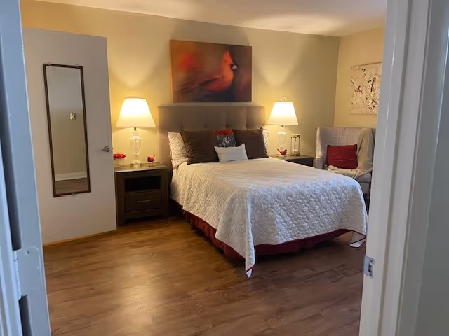 A cozy bedroom with a queen-sized bed covered in a white quilt and multiple pillows. Two bedside tables each have a lamp with a clear glass base. Above the bed hangs a painting of a red cardinal bird. To the right of the bed is a cushioned armchair with a red pillow. The floor is wooden, and a full-length mirror is mounted on the left wall near the door.