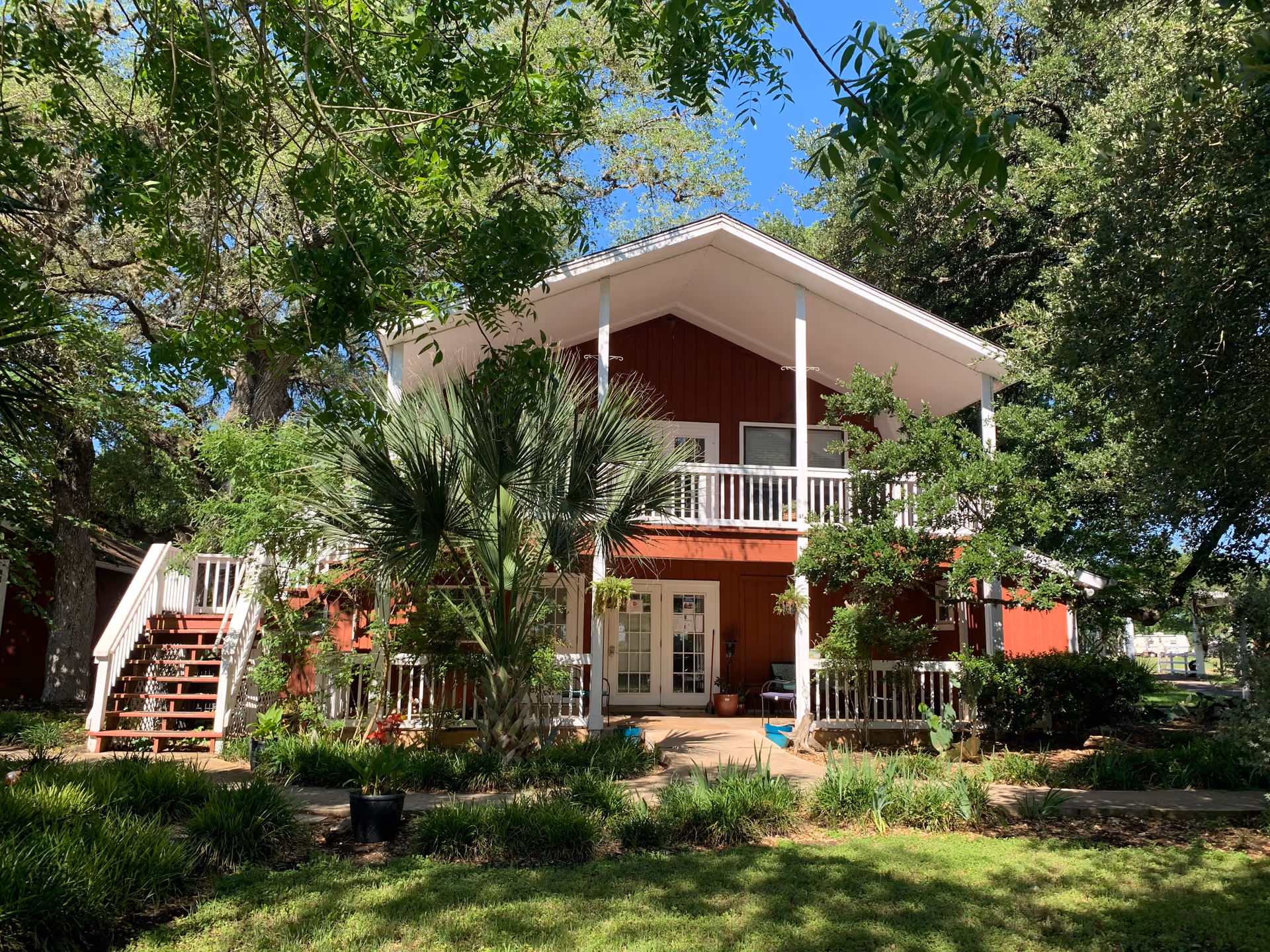 A two-story red building with white trim surrounded by lush green trees and plants under a clear blue sky. The building has a porch on the ground floor and a balcony on the upper floor, both with white railings. There is an outdoor staircase on the left side leading to the upper balcony.