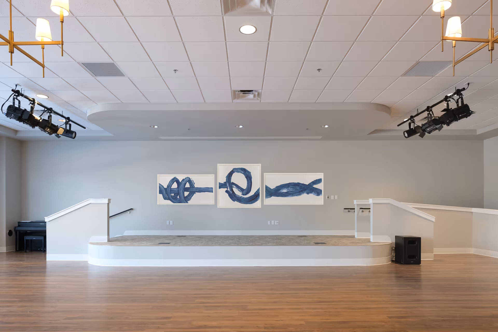 An empty stage area in a senior living facility with three abstract blue paintings on the wall behind it. The stage has carpeted flooring and is flanked by white railings with stairs on both sides. There are ceiling lights and stage spotlights above, a piano in the left corner, and a speaker on the right side on the wooden floor.