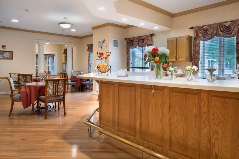 Interior view of a dining area in a senior living facility with wooden floors, tables covered with red tablecloths, and chairs with floral upholstery. A wooden counter with a white countertop is decorated with vases of flowers and a tiered fruit basket. Windows with patterned curtains allow natural light to enter the room.