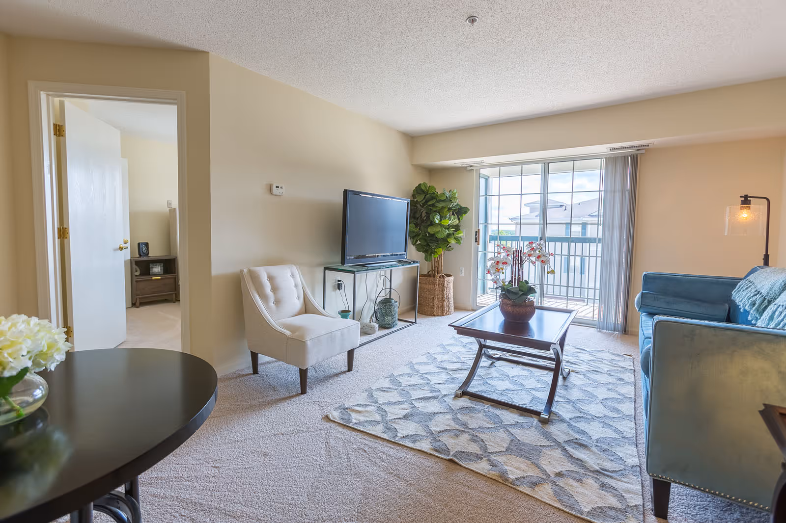 A bright living room with beige walls and carpet, featuring a blue sofa with a throw blanket, a white upholstered chair, a wooden coffee table with a potted orchid, a flat-screen TV on a glass stand, a large window with vertical blinds leading to a balcony, and a round table with a vase of white flowers.