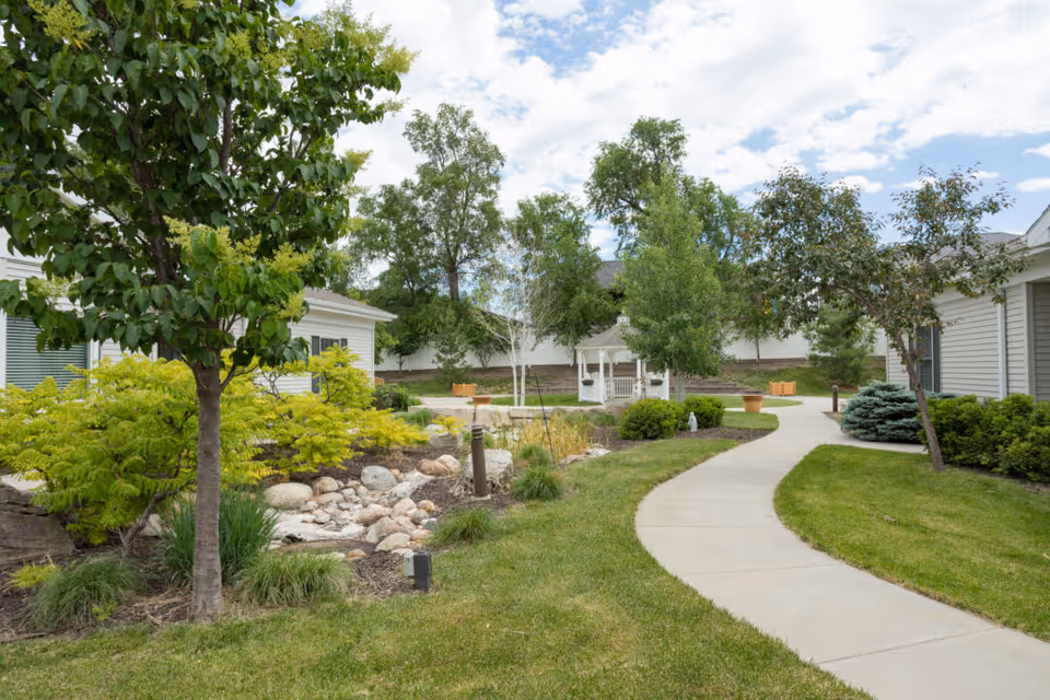 A landscaped outdoor garden area at CountryHouse Residence featuring a curved concrete walkway, green grass, various trees and shrubs, a small rock garden, and a white gazebo in the background under a partly cloudy sky.