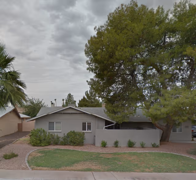 Grey single-story building with a large tree, shrubs, and a low front wall.