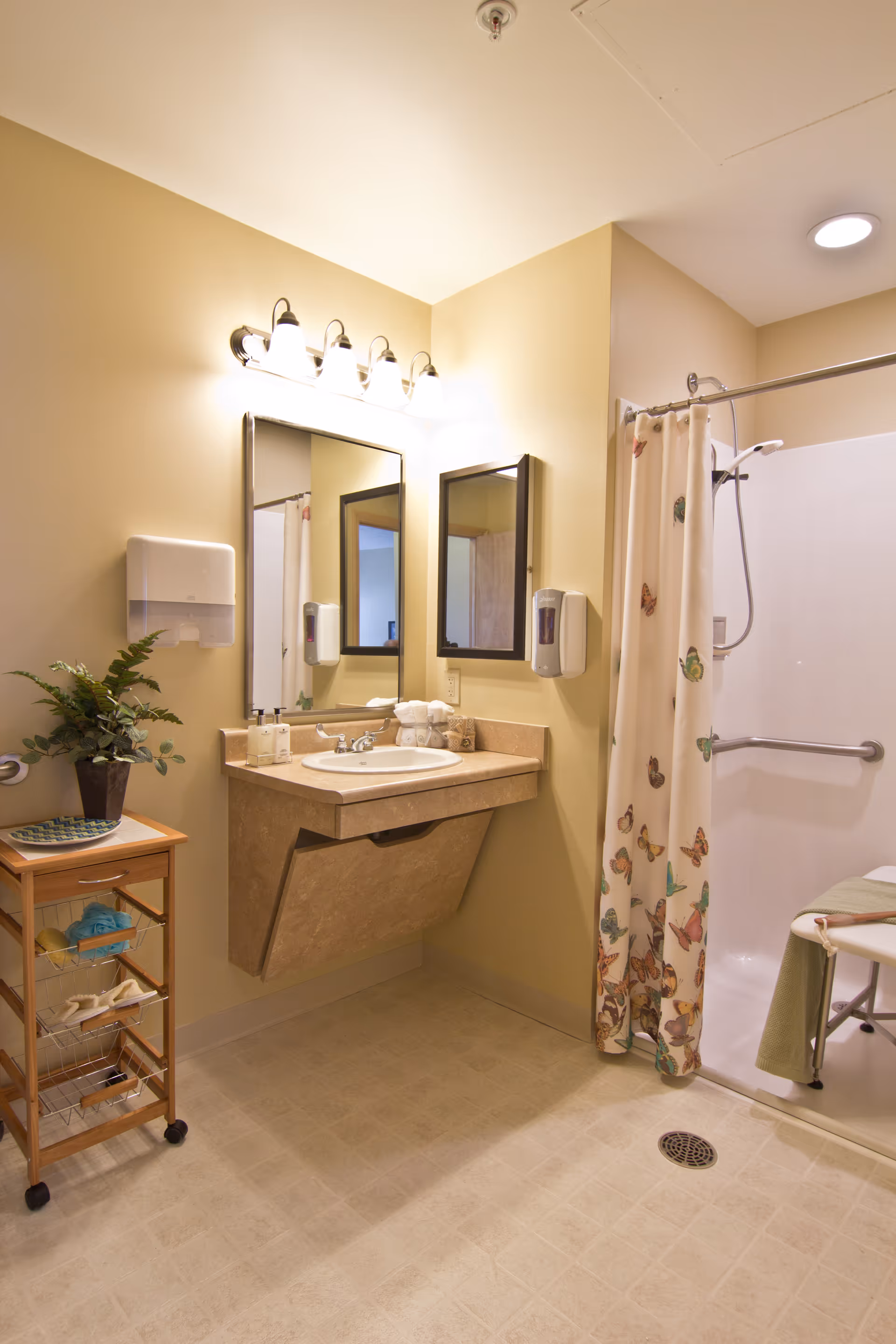 A clean and accessible bathroom with a beige color scheme. The image shows a wall-mounted sink with a large mirror above it, a soap dispenser, and a paper towel dispenser. To the right, there is a shower area with a curtain decorated with colorful butterflies and a shower chair inside. A small wooden cart with shelves and a potted plant on top is placed to the left of the sink.