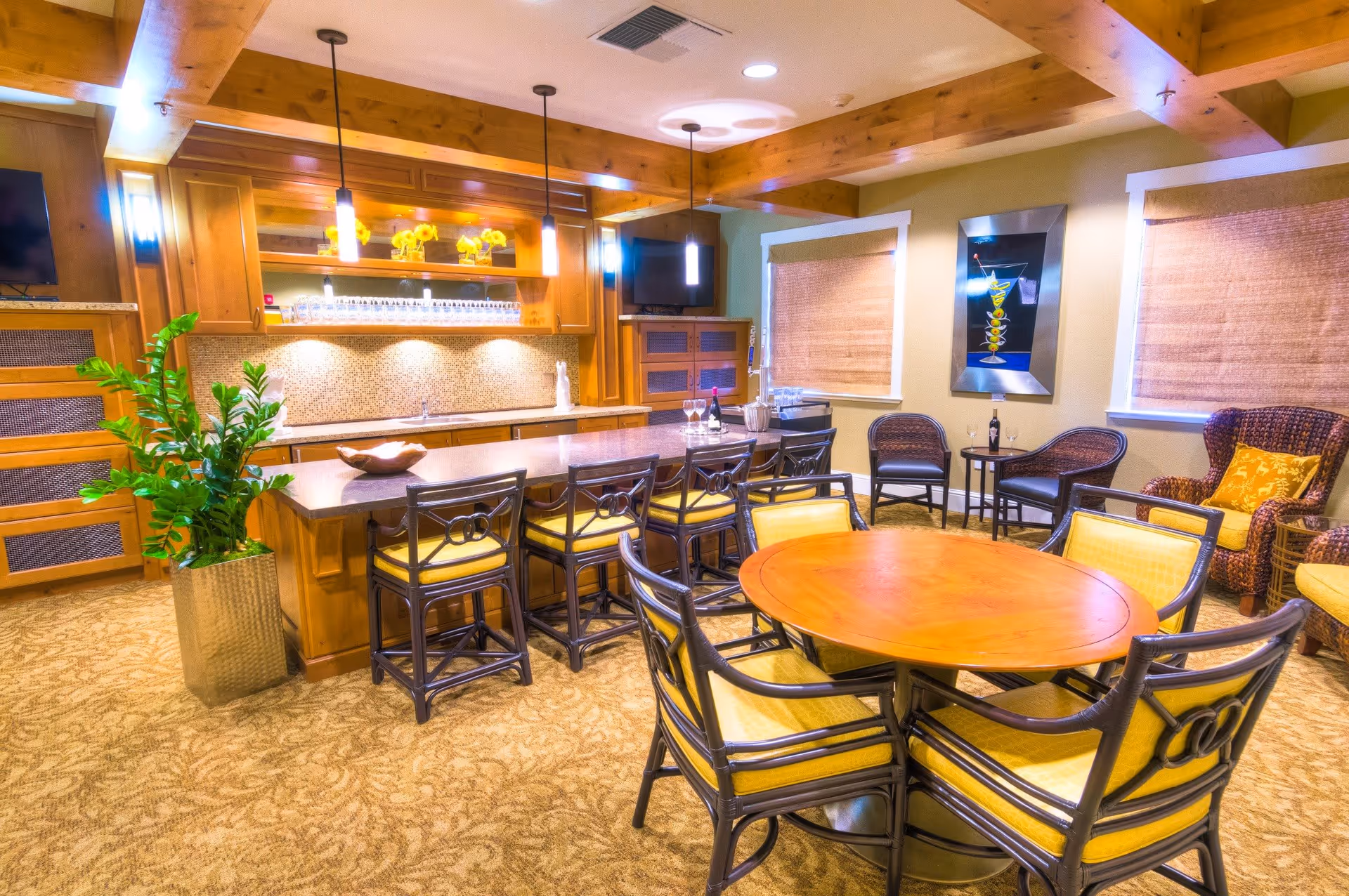 A cozy interior room with a wooden ceiling and warm lighting. The room features a large kitchen island with six chairs, a round wooden table with six chairs, and a seating area with wicker chairs and a small table. There are two windows with woven shades, a potted plant, and a wall-mounted TV. The decor includes a framed artwork and yellow flowers on a shelf above the kitchen counter.