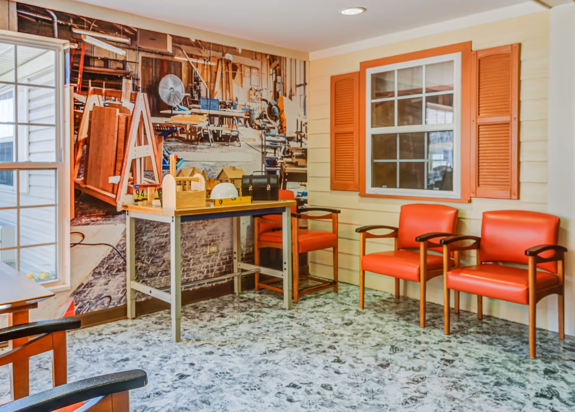 A room with three orange chairs with wooden armrests arranged against a wall with a window and orange shutters. A table with various wooden objects and a white hard hat is positioned near a wall mural depicting a workshop with tools and wood materials. The floor has a marbled pattern, and there is a large window on the left side letting in natural light.